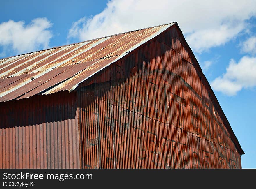 The metal roof and front of an old, rusty, metal building, stands against a blue sky with white clouds. The metal roof and front of an old, rusty, metal building, stands against a blue sky with white clouds.