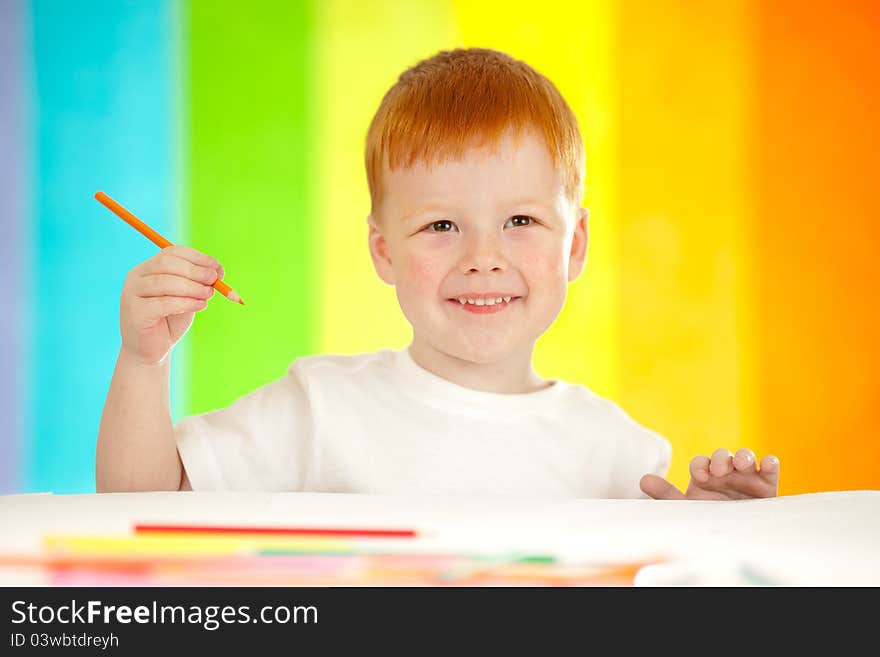 Red-haired adorable boy drawing with orange pencil on rainbow background. Red-haired adorable boy drawing with orange pencil on rainbow background