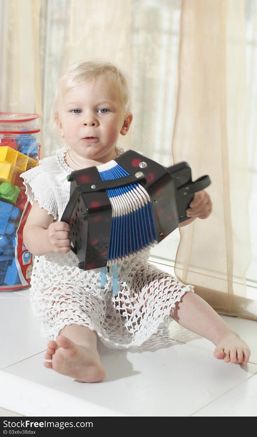 The little girl in a white dress sits on a window sill and plays an accordion