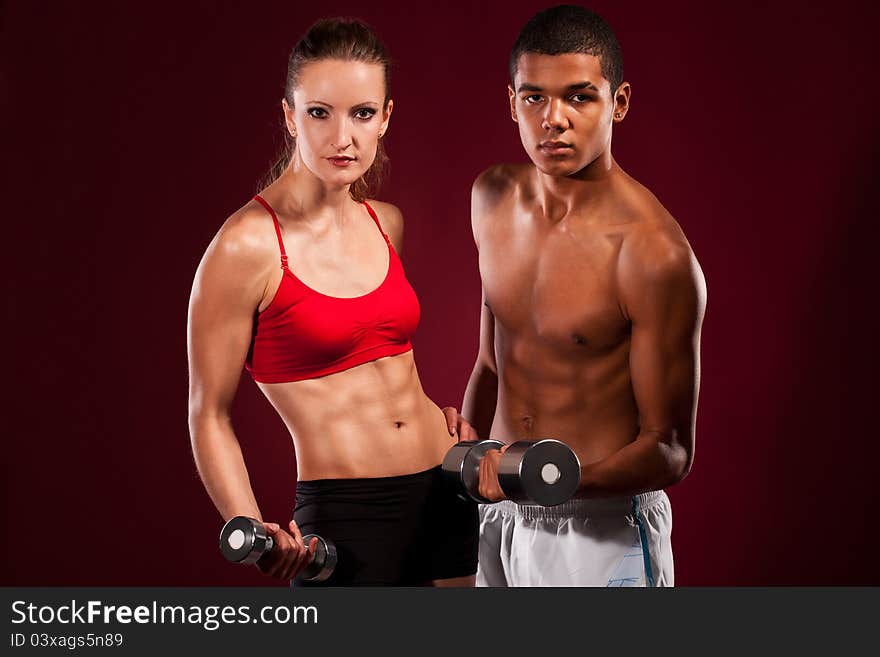 Strong young couple working out with dumbbells. Shot in studio on a red background.