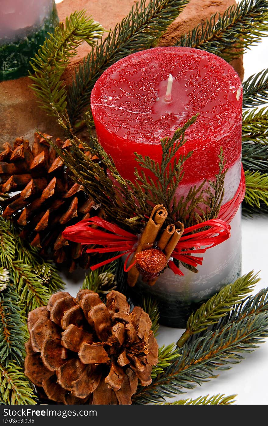 Red, white and green festive candle with green cedar leaves and cinnamon sticks tied on with twine for Christmas. The candle is placed among pine branches and pine cones. Red, white and green festive candle with green cedar leaves and cinnamon sticks tied on with twine for Christmas. The candle is placed among pine branches and pine cones.