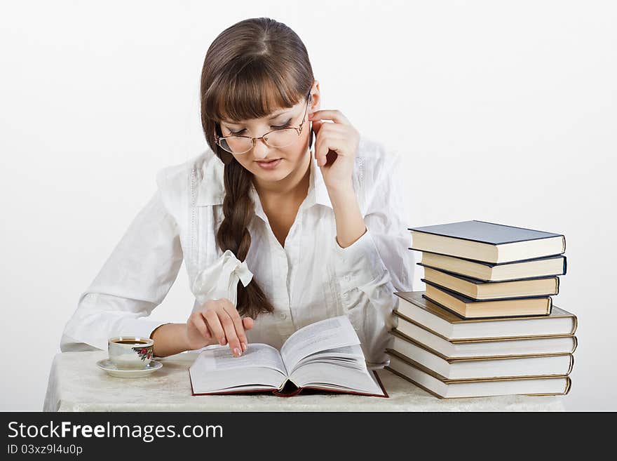 Beautiful girl with glasses in a white shirt thoughtfully reading a book at the table. By her side there is a pile of books and a cup of coffee. Her Hair is braided into a braid. Photographed in a studio against a white background. Beautiful girl with glasses in a white shirt thoughtfully reading a book at the table. By her side there is a pile of books and a cup of coffee. Her Hair is braided into a braid. Photographed in a studio against a white background.