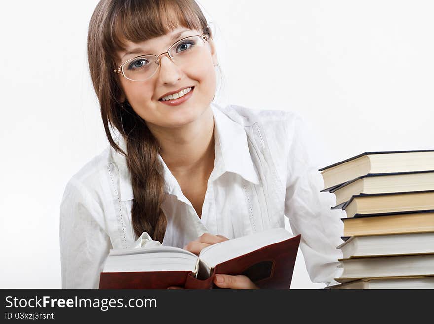 Beautiful girl with glasses in a white shirt reading a book at the table. The girl looks into the camera and sweet smiles. Her Hair is braided into a braid. Fragmant of a stack of books can be seen near the edge of photo. Photographed in a studio against a white background. Beautiful girl with glasses in a white shirt reading a book at the table. The girl looks into the camera and sweet smiles. Her Hair is braided into a braid. Fragmant of a stack of books can be seen near the edge of photo. Photographed in a studio against a white background.