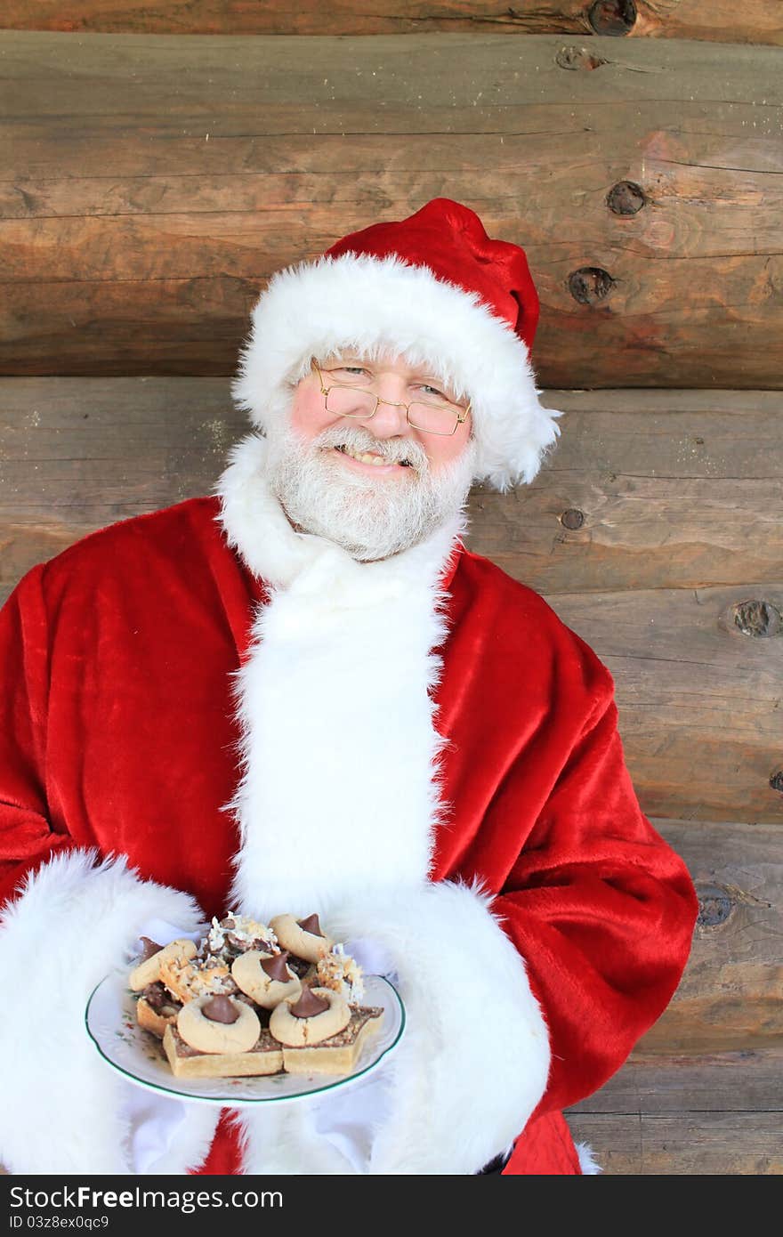 Santa standing in front of a log cabin wall holding a plate of Christmas cookies. Santa standing in front of a log cabin wall holding a plate of Christmas cookies.