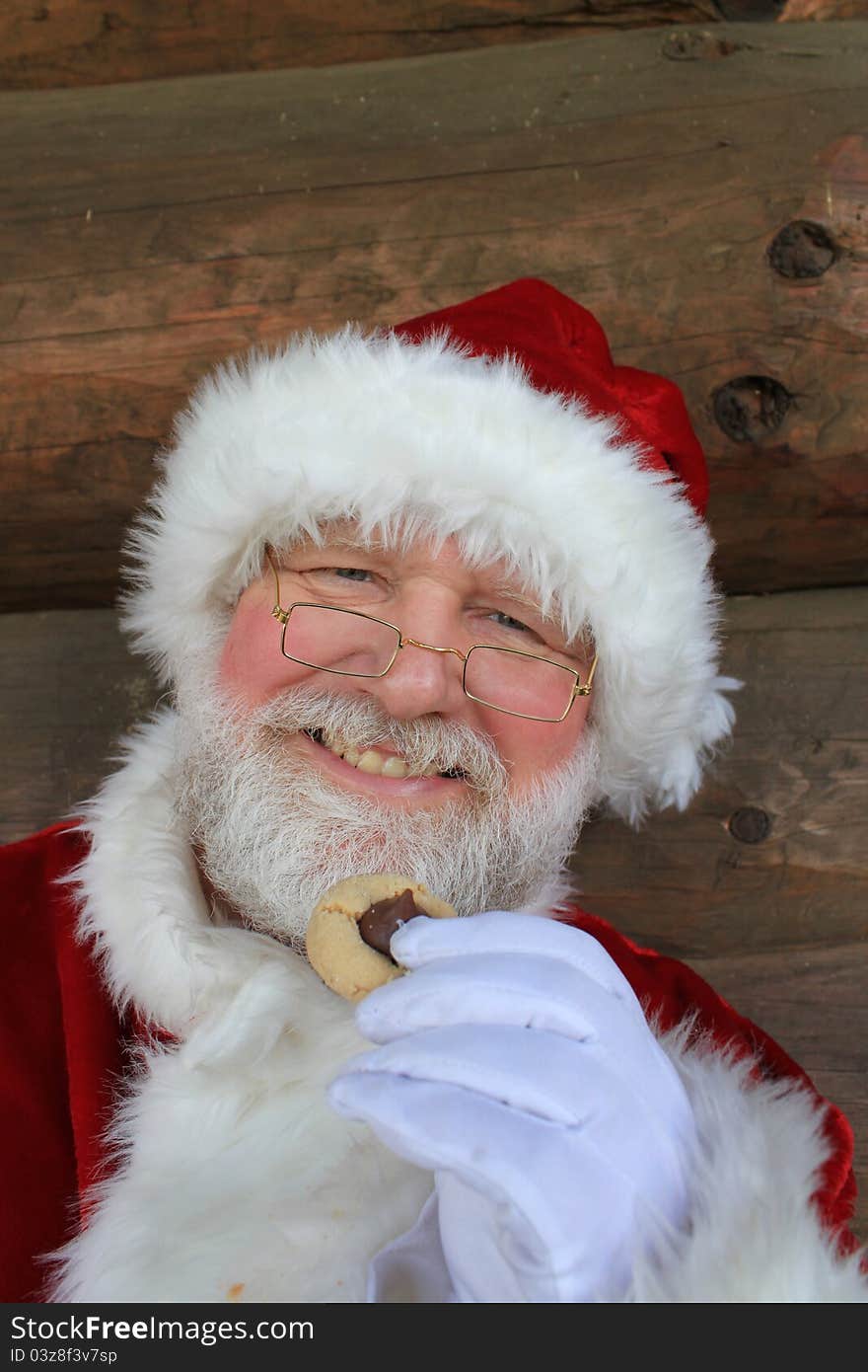 Santa Claus eating a traditional Christmas cookie in front of the wall of a log cabin. Santa Claus eating a traditional Christmas cookie in front of the wall of a log cabin.