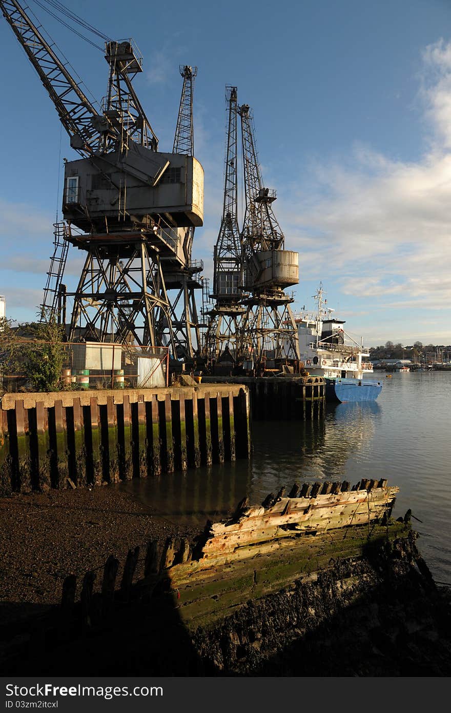 Old dockyard cranes on a harbour side jetty with an old rotting hulk in the foreground. Cloudy blue sky with moody lighting and shadows. River Itchen. Southampton. England. Old dockyard cranes on a harbour side jetty with an old rotting hulk in the foreground. Cloudy blue sky with moody lighting and shadows. River Itchen. Southampton. England.