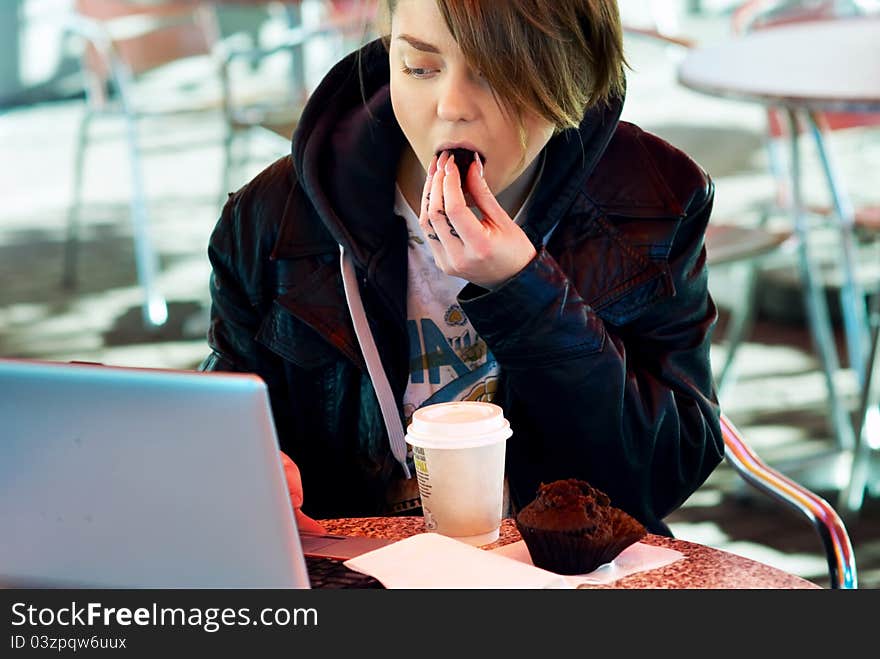 Portrait of blond stylish girl with cup and muffin on the terrace of the fast food with laptop on the table