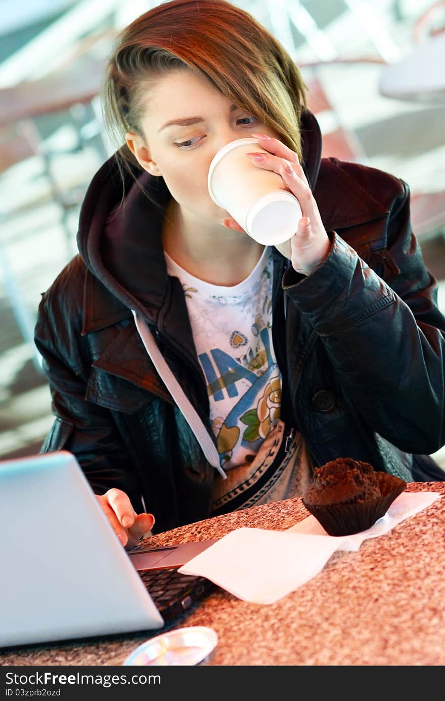 Portrait of blond stylish girl with cup and muffin on the terrace of the fast food with laptop on the table