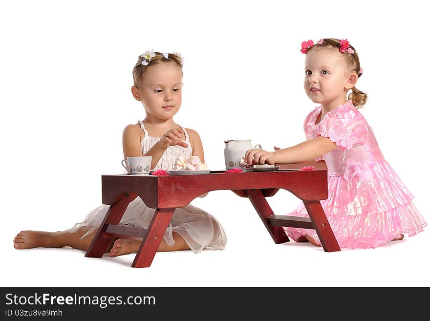 Two pretty twins sisters in beautiful dresses having tea wtih zephyr sitting at coffee table isolated on white background. Two pretty twins sisters in beautiful dresses having tea wtih zephyr sitting at coffee table isolated on white background