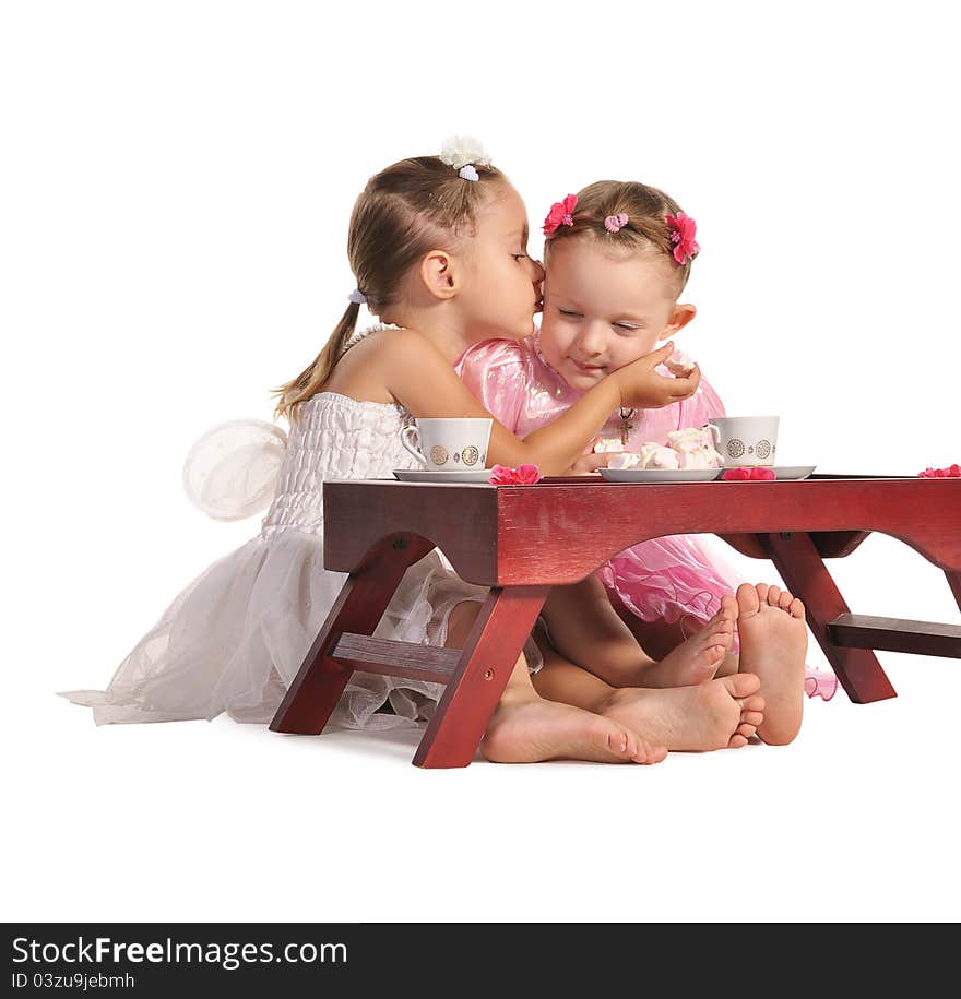 Two pretty twins sisters in beautiful dresses having tea wtih zephyr sitting at coffee table isolated on white background. Two pretty twins sisters in beautiful dresses having tea wtih zephyr sitting at coffee table isolated on white background