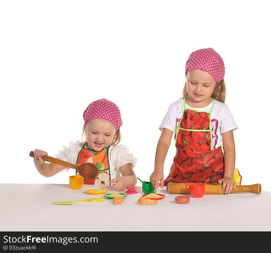 Two pretty little twins sisters wearing spotted pink headscarfs and colourful aprons pretending housewifes cooking isolated on white background. Two pretty little twins sisters wearing spotted pink headscarfs and colourful aprons pretending housewifes cooking isolated on white background