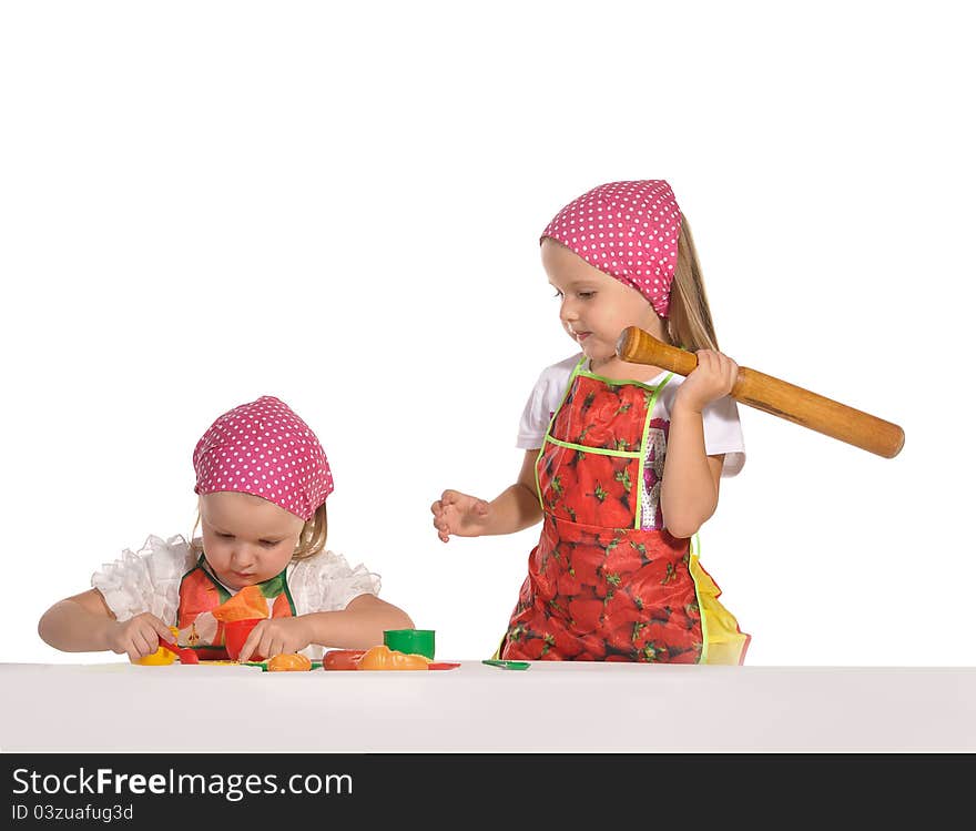 Two pretty little twins sisters wearing spotted pink headscarfs and colourful aprons pretending housewifes cooking isolated on white background. Two pretty little twins sisters wearing spotted pink headscarfs and colourful aprons pretending housewifes cooking isolated on white background