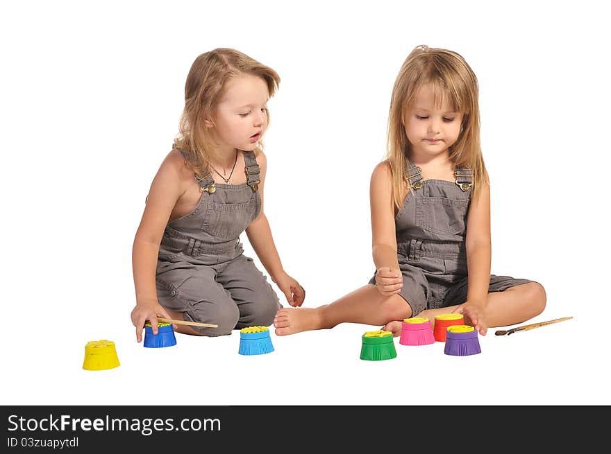 Two pretty barefooted twins sisters in rompers choosing paints isolated on white background. Two pretty barefooted twins sisters in rompers choosing paints isolated on white background