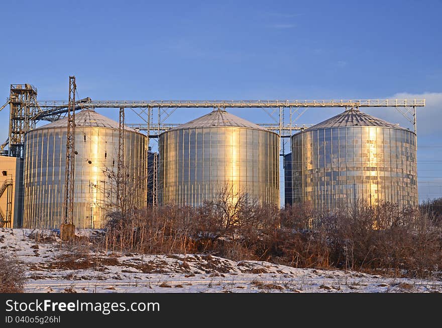 Group of silos filled with cereal grain against blue sky, placed in ploughed acres