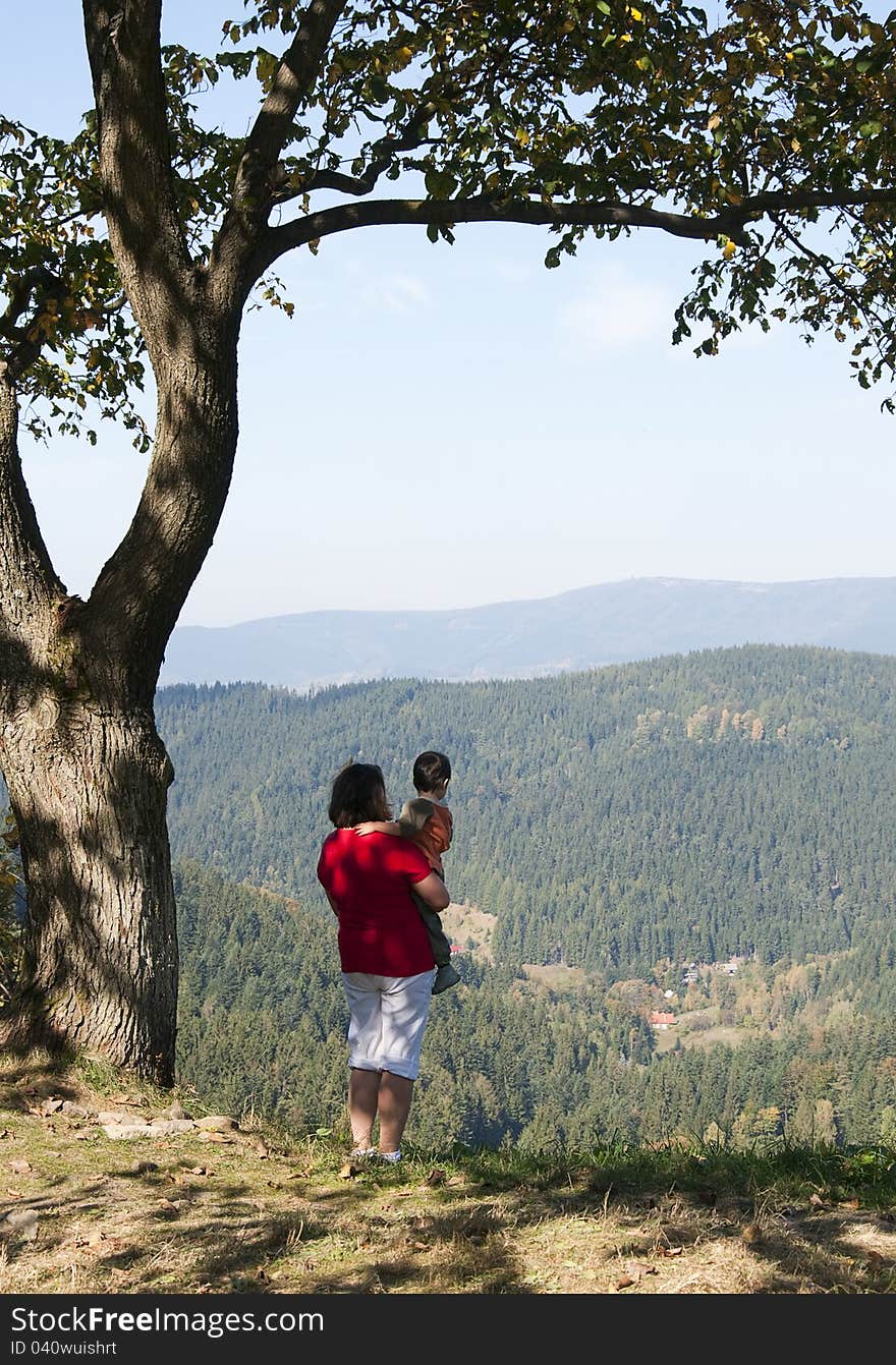 Mother and child standing on the top of a hill under a tree looking down at the green forest. Mother and child standing on the top of a hill under a tree looking down at the green forest.