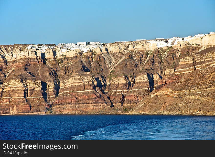 A high stone wall overlooking the sea. This stone stands in front of the island of Santorini travelers. Its shores are steep and precipitous. On top of a mountain visible white city. A high stone wall overlooking the sea. This stone stands in front of the island of Santorini travelers. Its shores are steep and precipitous. On top of a mountain visible white city.