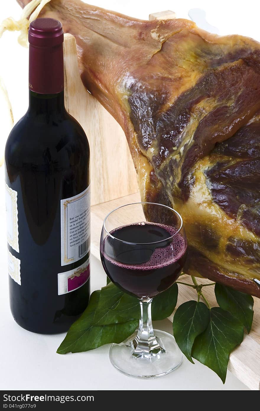 A front leg Serrano ham on displayed on a wooden rack and a bottle of red wine with a full glass of wine on a white background. A front leg Serrano ham on displayed on a wooden rack and a bottle of red wine with a full glass of wine on a white background.