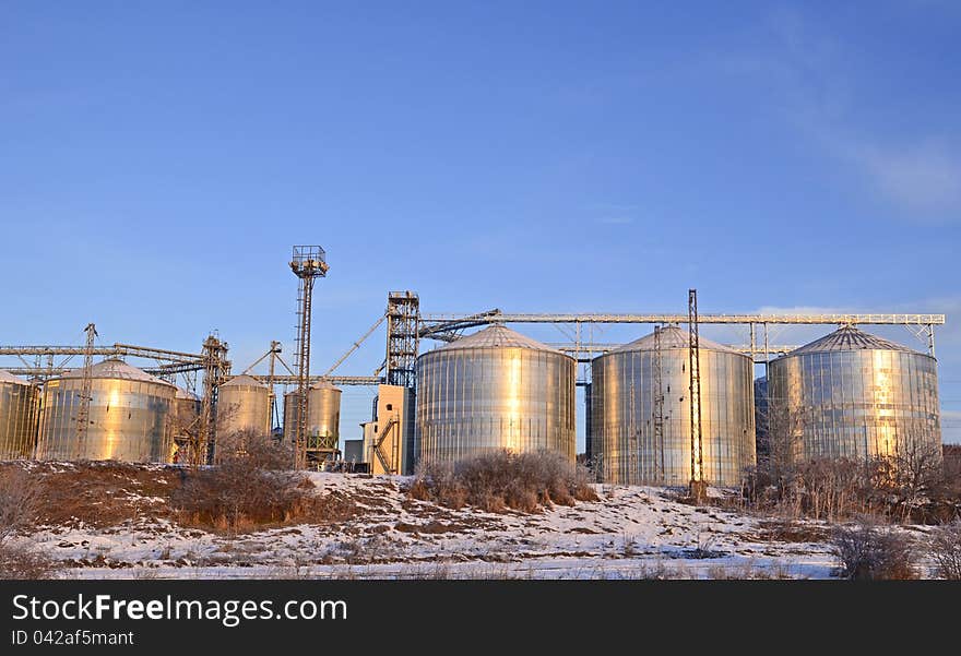 Group of silos filled with cereal grain against blue sky, placed in ploughed acres