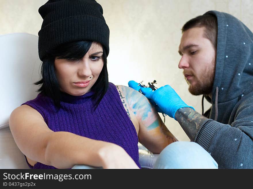 Emotions of a girl while a tattoo artist applying his craft on her shoulder at his workshop. Focus on female's face. Emotions of a girl while a tattoo artist applying his craft on her shoulder at his workshop. Focus on female's face.