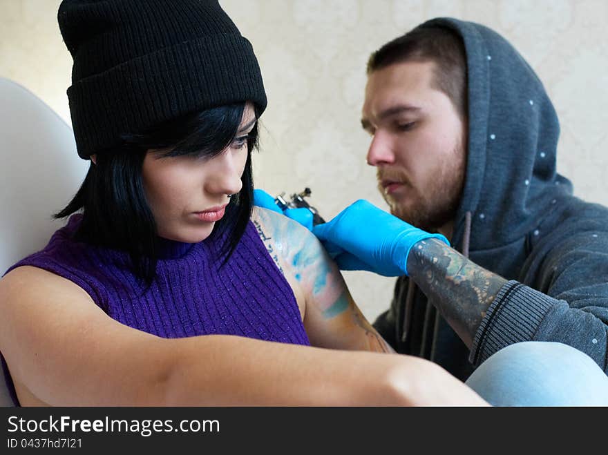 Emotions of a girl while a tattoo artist applying his craft on her shoulder at his workshop. Focus on female's face. Emotions of a girl while a tattoo artist applying his craft on her shoulder at his workshop. Focus on female's face.