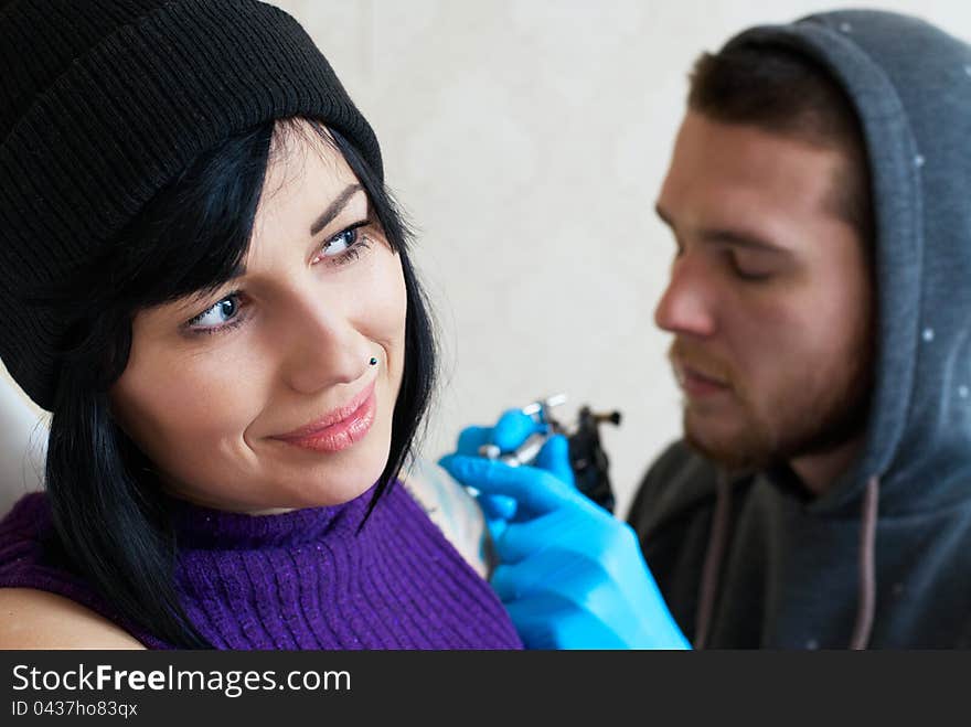 Emotions of a girl while a tattoo artist applying his craft on her shoulder at his workshop. Focus on female's face. Emotions of a girl while a tattoo artist applying his craft on her shoulder at his workshop. Focus on female's face.