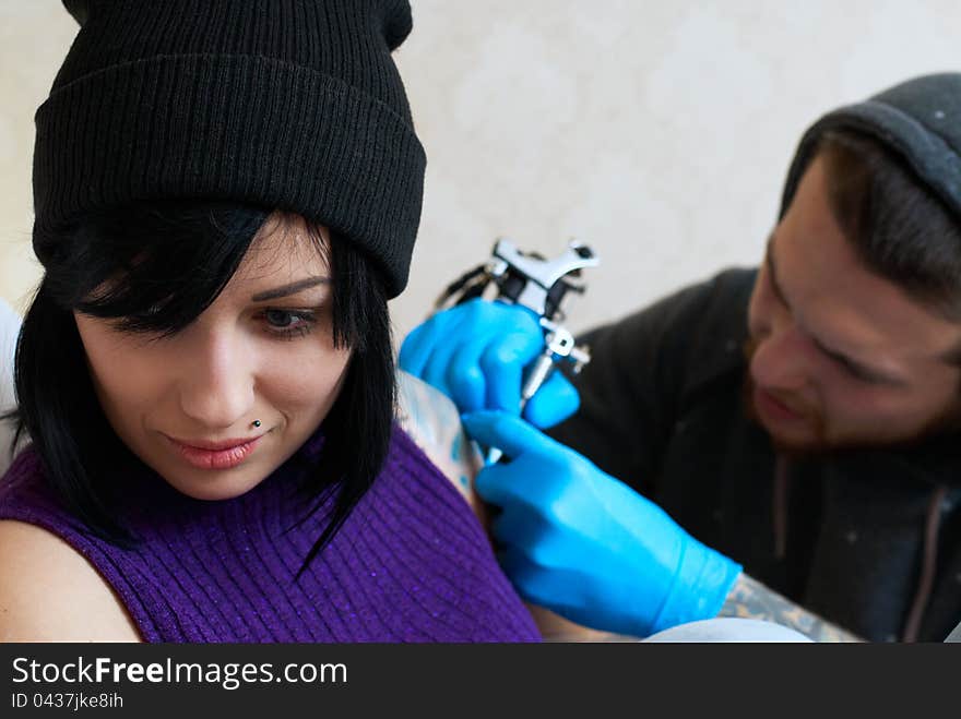 Emotions of a girl while a tattoo artist applying his craft on her shoulder at his workshop. Focus on female's face. Emotions of a girl while a tattoo artist applying his craft on her shoulder at his workshop. Focus on female's face.