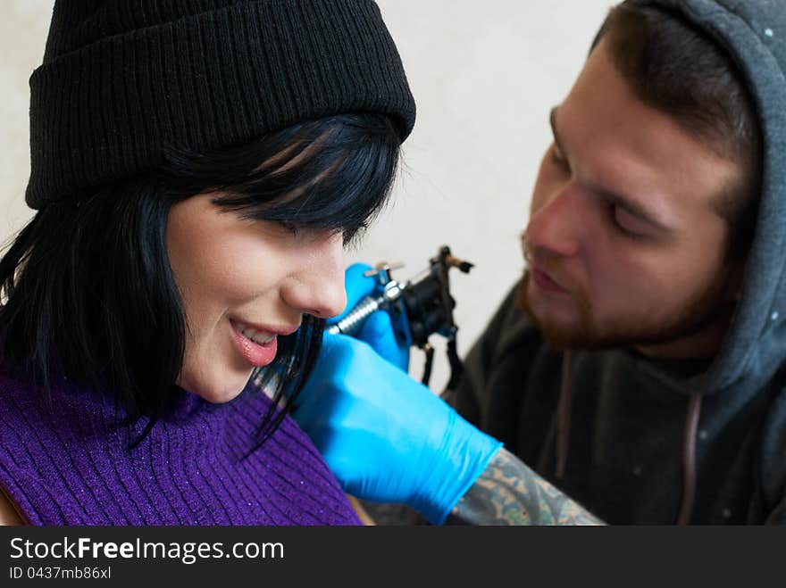 Emotions of a girl while a tattoo artist applying his craft on her shoulder at his workshop. Focus on female's face. Emotions of a girl while a tattoo artist applying his craft on her shoulder at his workshop. Focus on female's face.