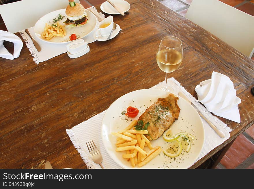 Fried hake fish in batter and chips on a white plate with a glass of chilled white wine and a hamburger and chips in a gourmet restaurant. Fried hake fish in batter and chips on a white plate with a glass of chilled white wine and a hamburger and chips in a gourmet restaurant