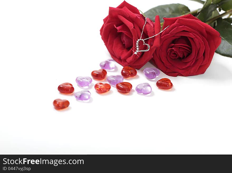 A still life composition of a heart-shaped white gold and diamond necklace hanging from two red roses with heart-shaped acrylic decors on the foreground with lots of white copy space. A still life composition of a heart-shaped white gold and diamond necklace hanging from two red roses with heart-shaped acrylic decors on the foreground with lots of white copy space