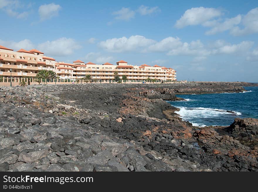 Image shows a hotel at the coast of Fuerteventura. Stone coast is black, ocean and sky are blue, sky is light cloudy. Image shows a hotel at the coast of Fuerteventura. Stone coast is black, ocean and sky are blue, sky is light cloudy.