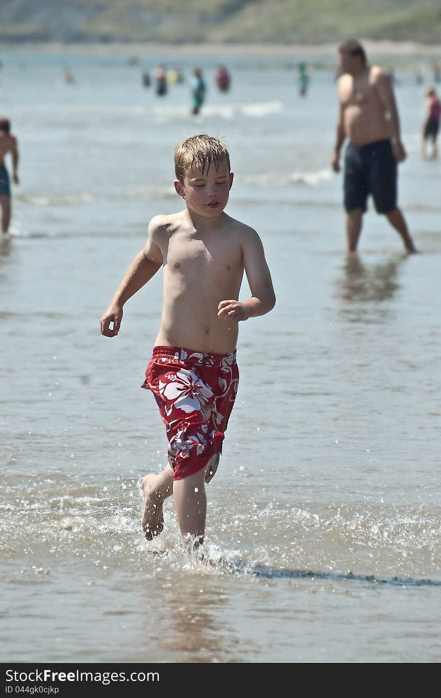 An image of a young boy wearing boardshorts running through the water on a hot summers day. An image of a young boy wearing boardshorts running through the water on a hot summers day