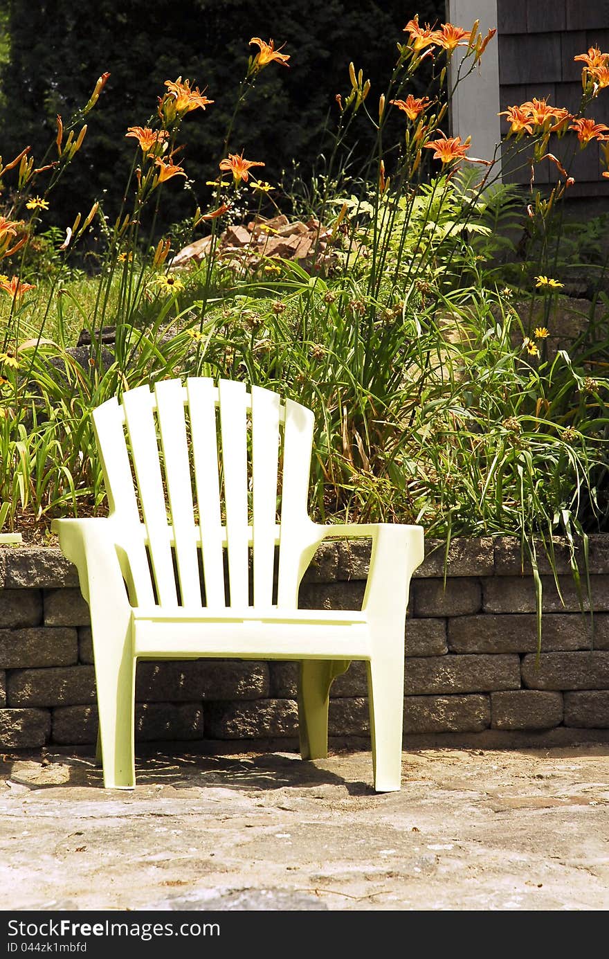 A white garden chair stands on a patio in front of a stone wall and tiger lilies. A white garden chair stands on a patio in front of a stone wall and tiger lilies