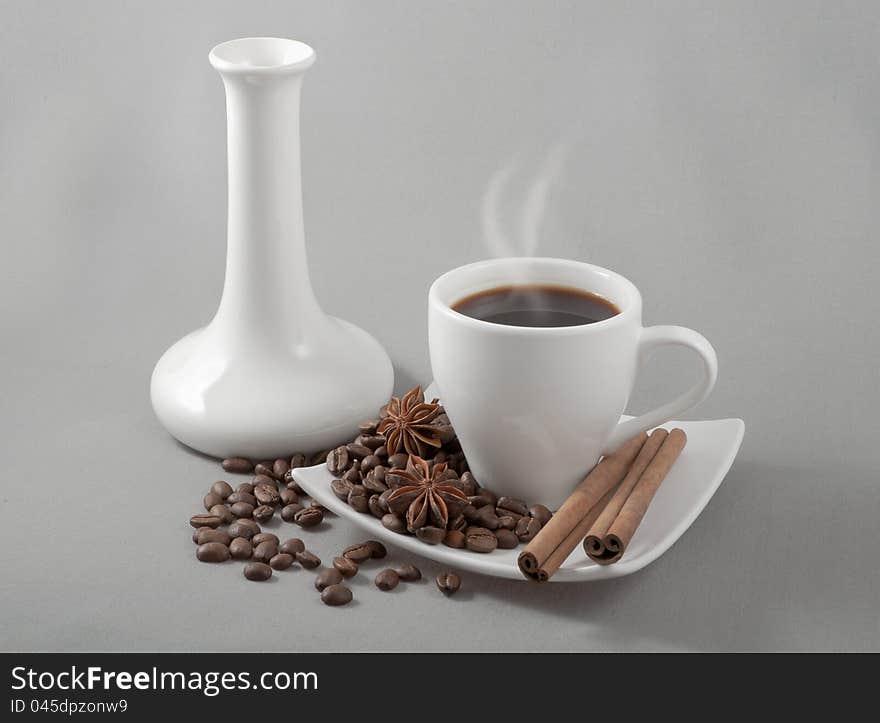 Black coffee in a white mug stands on a saucer with the spilled grains on a background a white vase. Black coffee in a white mug stands on a saucer with the spilled grains on a background a white vase