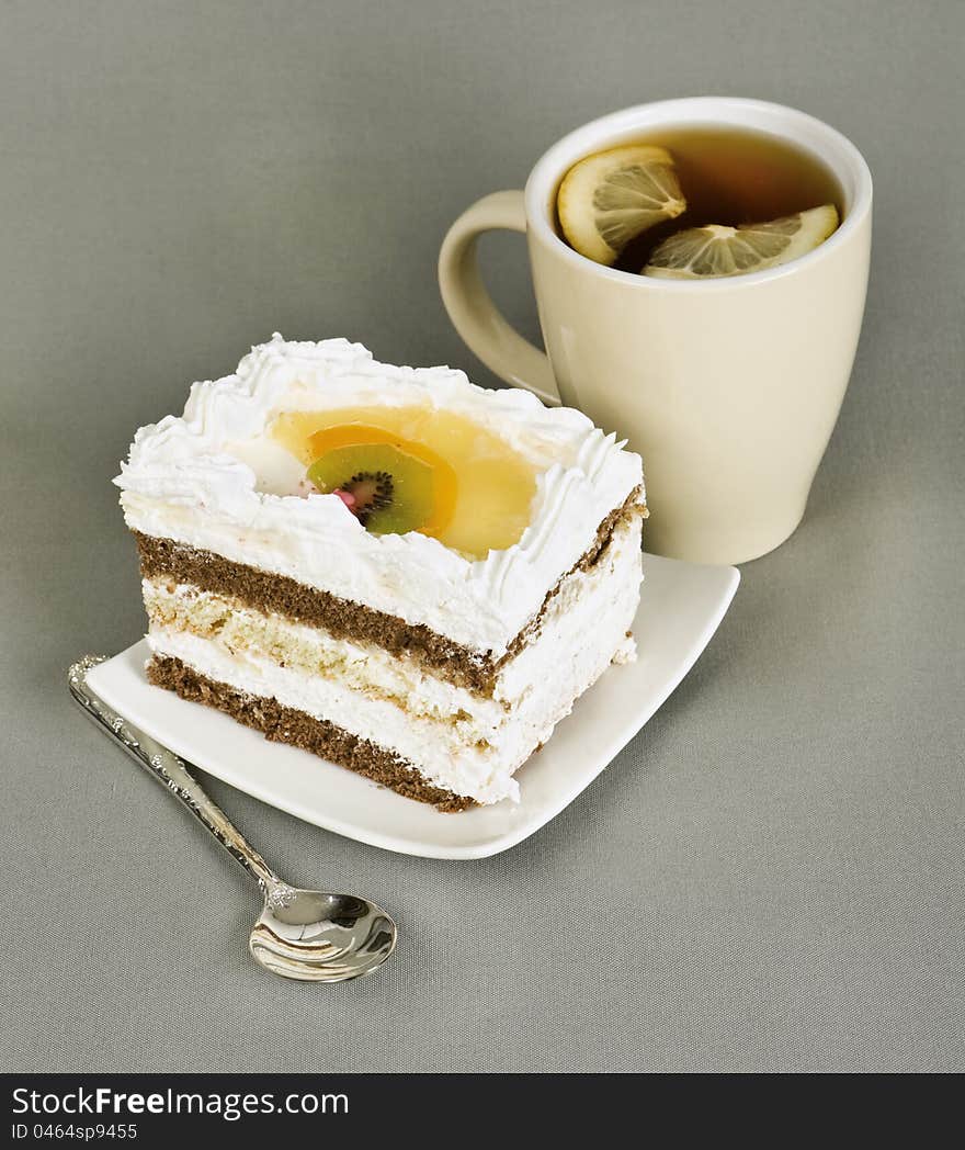 Piece of cake with fruit, tea with a lemon, tea-spoon on grey fabric. Piece of cake with fruit, tea with a lemon, tea-spoon on grey fabric