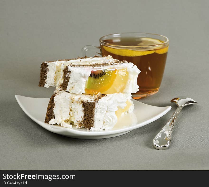 Piece of cake with fruit, tea with a lemon, tea-spoon on grey fabric. Piece of cake with fruit, tea with a lemon, tea-spoon on grey fabric