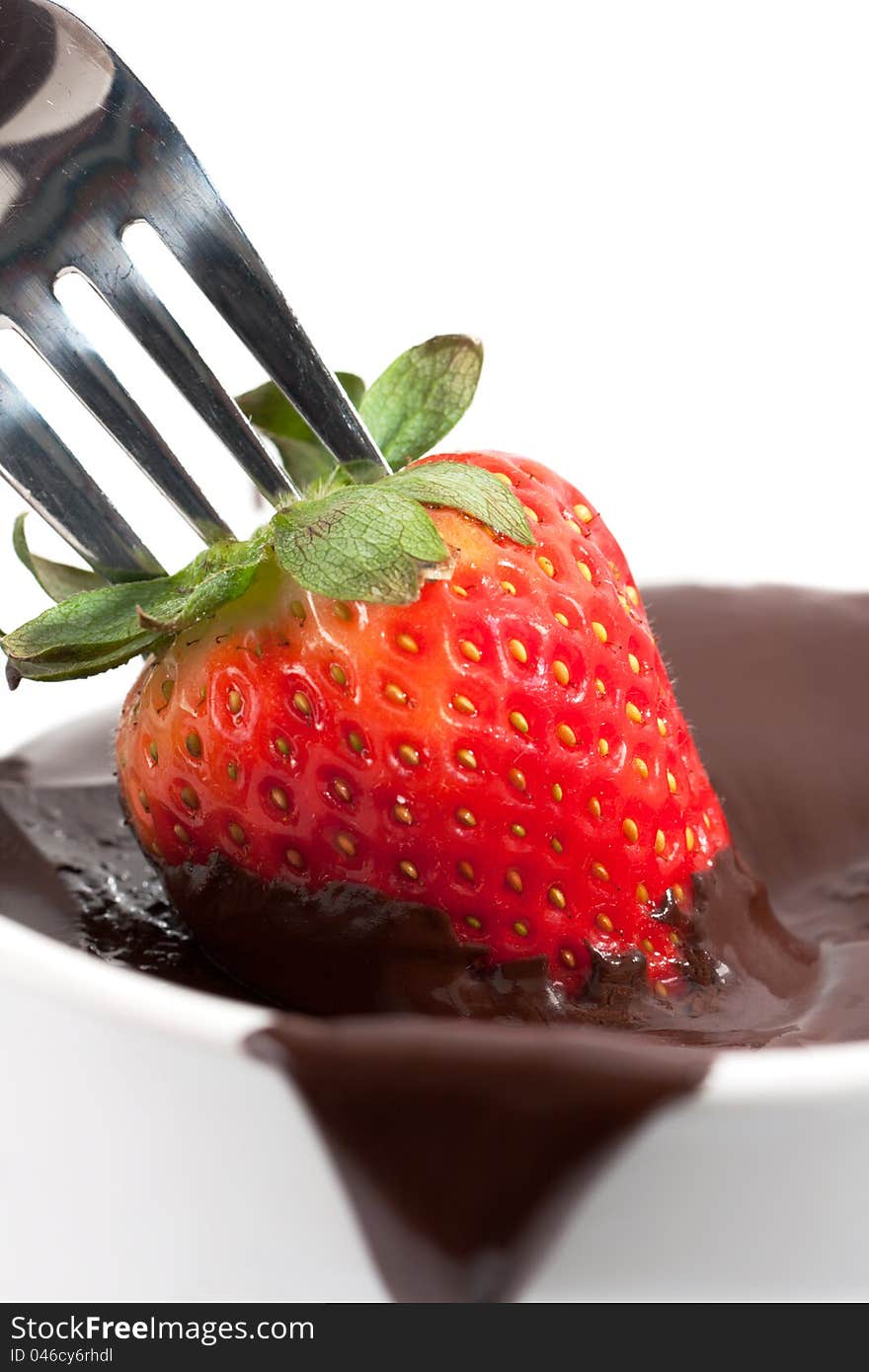 A ripe red strawberry on the end of a fork is being dipped into a bowl of melted brown chocolate. A ripe red strawberry on the end of a fork is being dipped into a bowl of melted brown chocolate