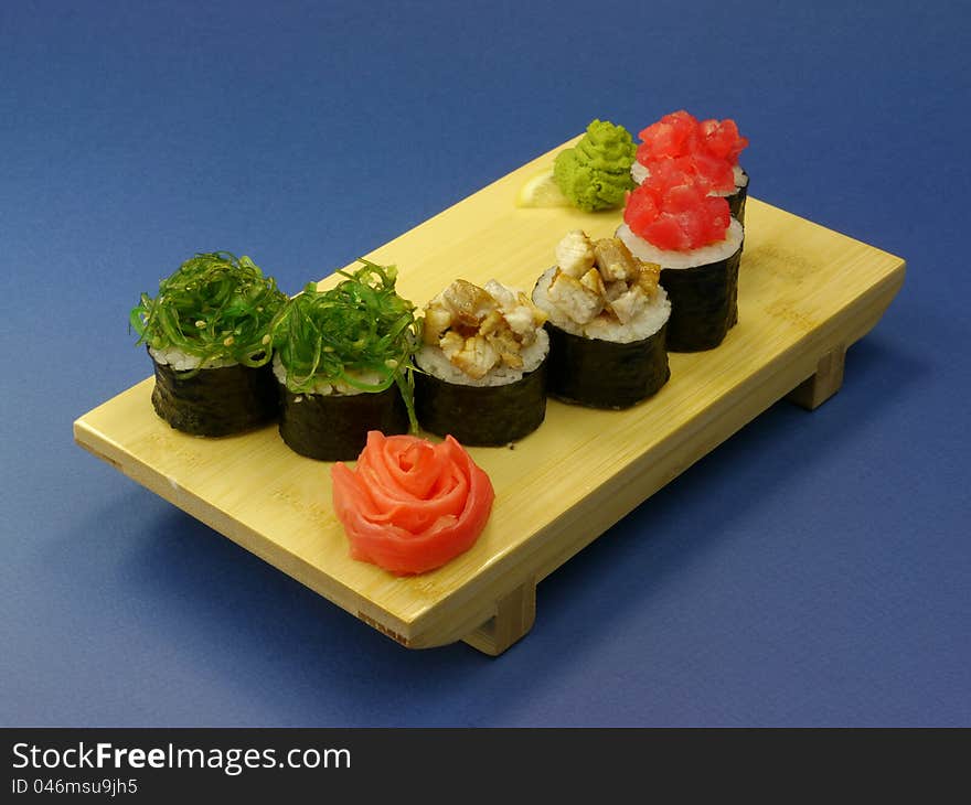 Delicious sushi rolls on wooden plate with pickled ginger and wasabi on blue background.