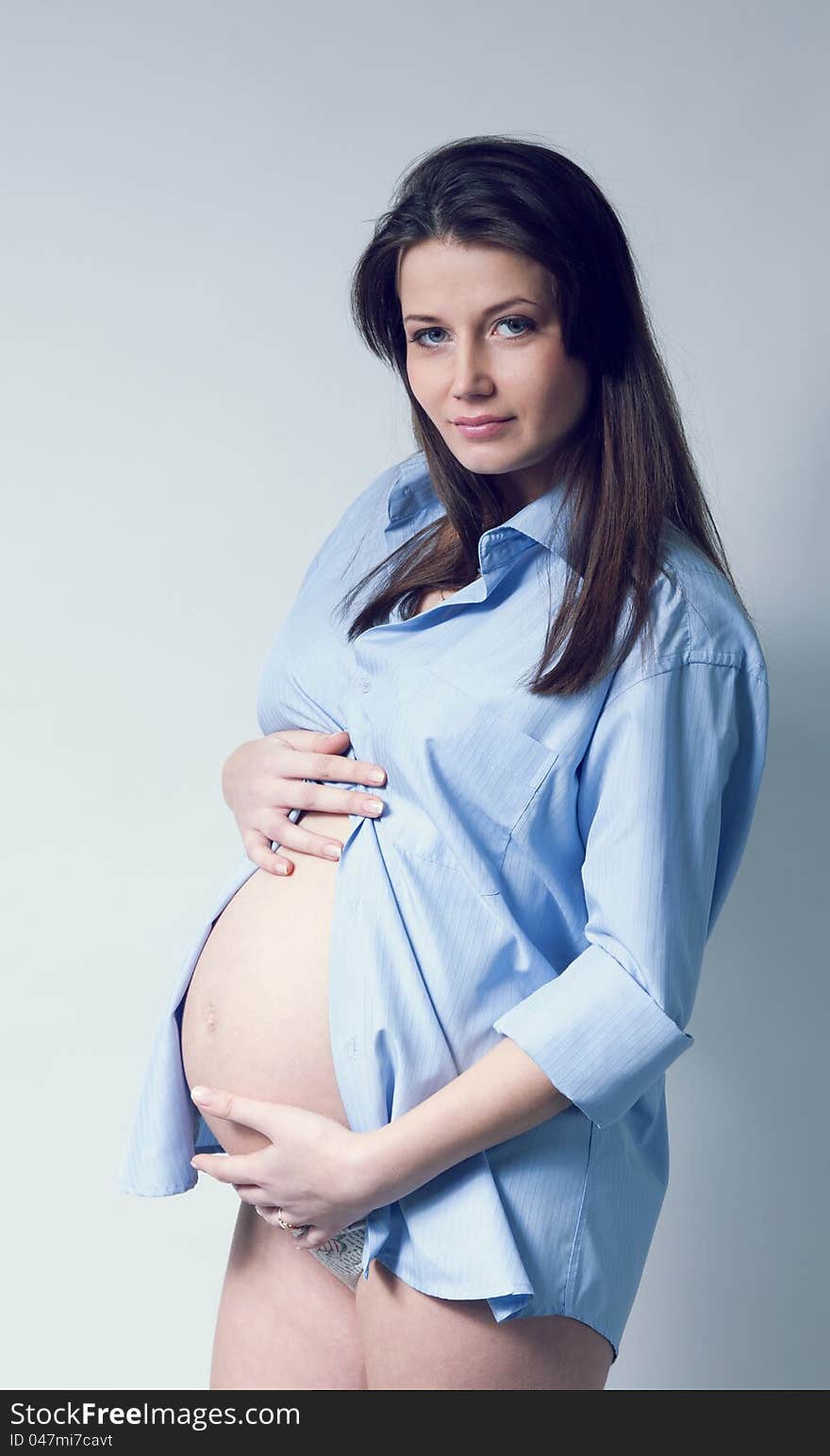 Beautiful pregnant white woman wearing blue shirt. Long dark hair. Hands on belly. Pregnancy collection.