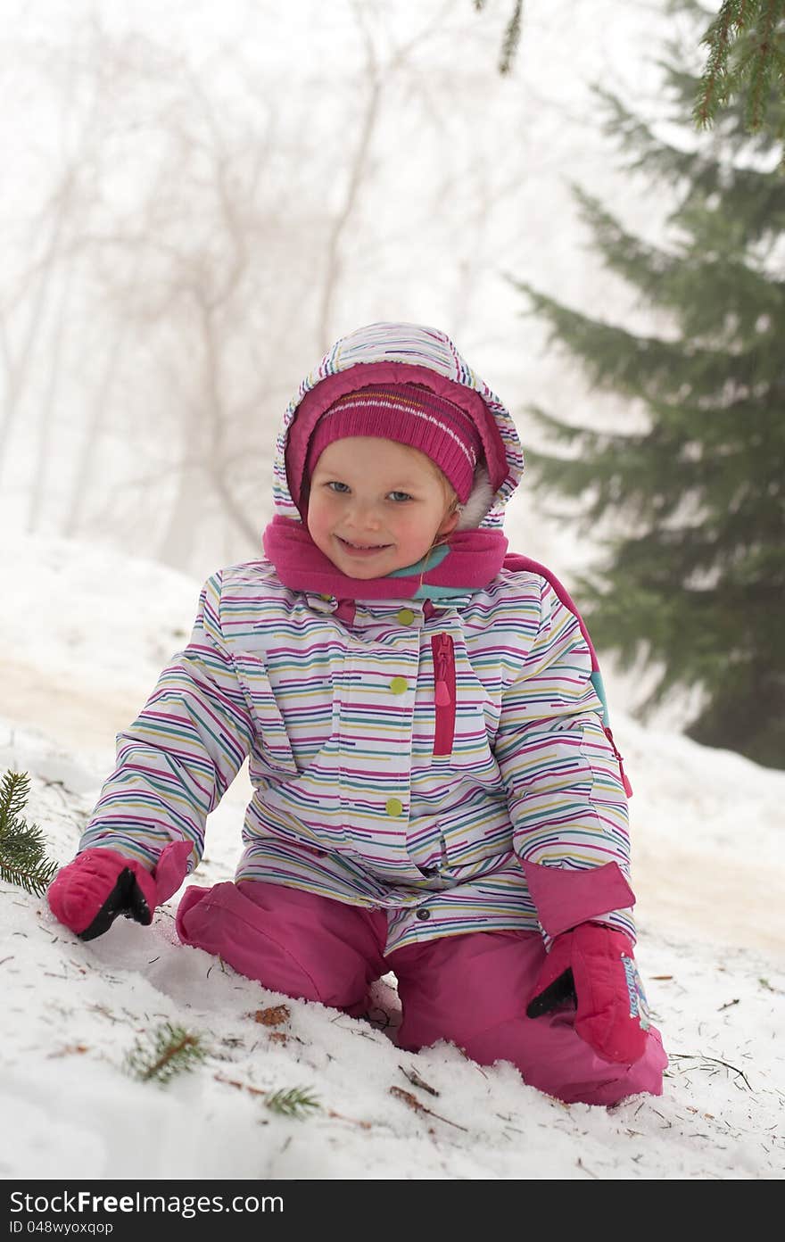 Girl sitting on the snow in the park with trees in the background, dressed in winter snow suite. Girl sitting on the snow in the park with trees in the background, dressed in winter snow suite