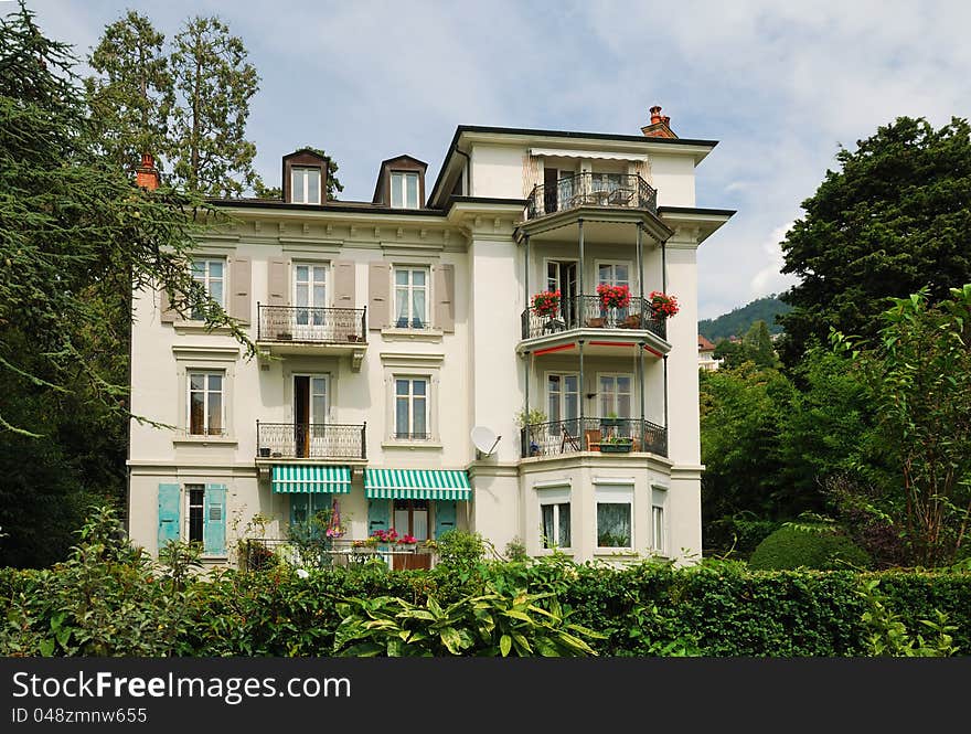 Detached house with shutters and hedge is in the middle of green trees and bushes. It is decorated with flowers. Detached house with shutters and hedge is in the middle of green trees and bushes. It is decorated with flowers