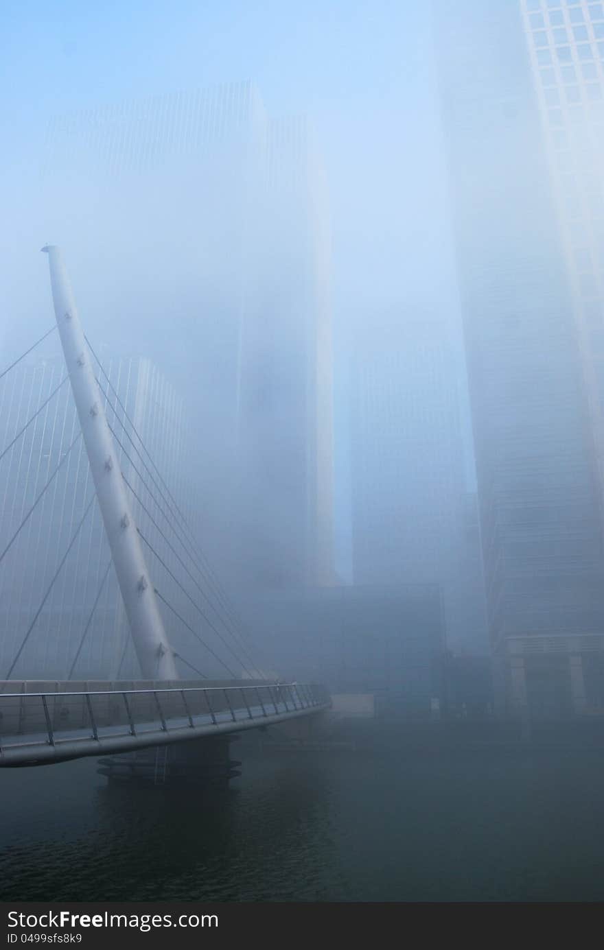 Thick fog obscures the view towards a bright and sunny Canary Wharf, seen from West India Dock, beside the South Quay Footbridge, London Docklands. Modern suspension bridge and modern skyscraper towers emerge from the sun and the mist. . Thick fog obscures the view towards a bright and sunny Canary Wharf, seen from West India Dock, beside the South Quay Footbridge, London Docklands. Modern suspension bridge and modern skyscraper towers emerge from the sun and the mist.