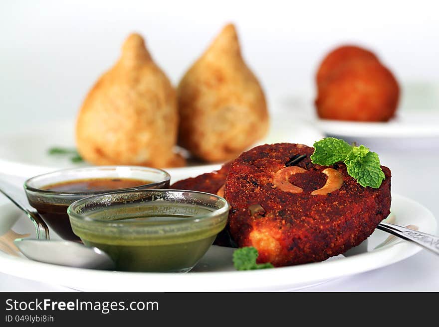 Indian deep fried snack cutlet made of potatoes, masala and cashew with chutneys and another popular snack samosa in the background