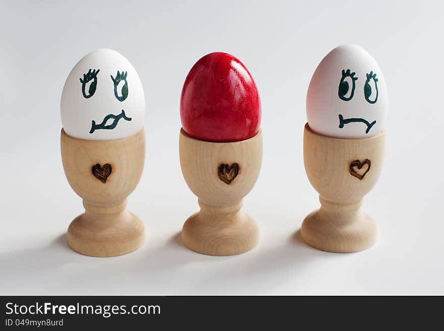 Three eggs in a egg cup, standing in a row. The egg in the middle is a red colored easter egg and the left and right one are looking surprised two the easter egg. Three eggs in a egg cup, standing in a row. The egg in the middle is a red colored easter egg and the left and right one are looking surprised two the easter egg.