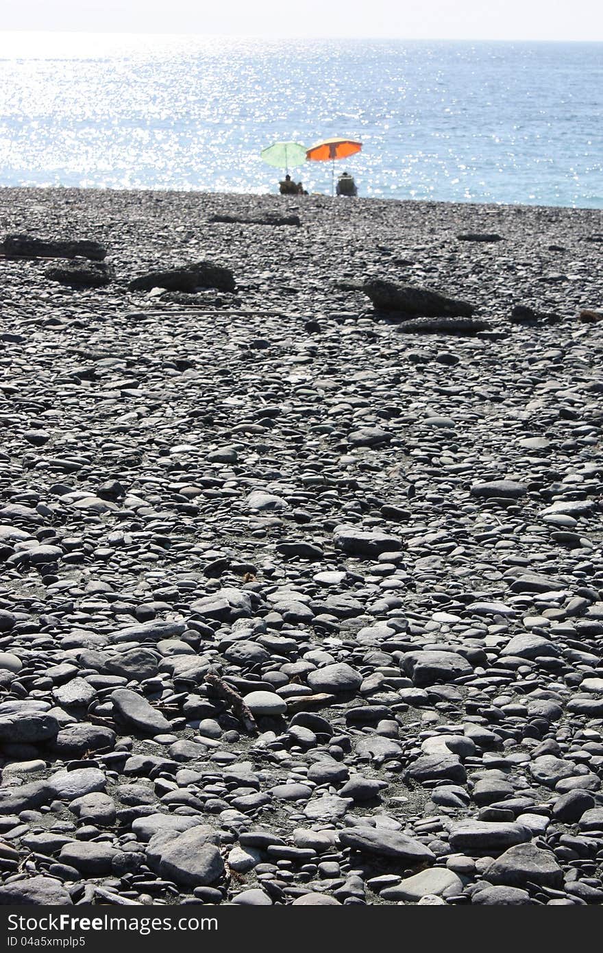 A black pebble beach. In the distance two colorfull umbrellas at the waters edge. (Nonza - Corsica)