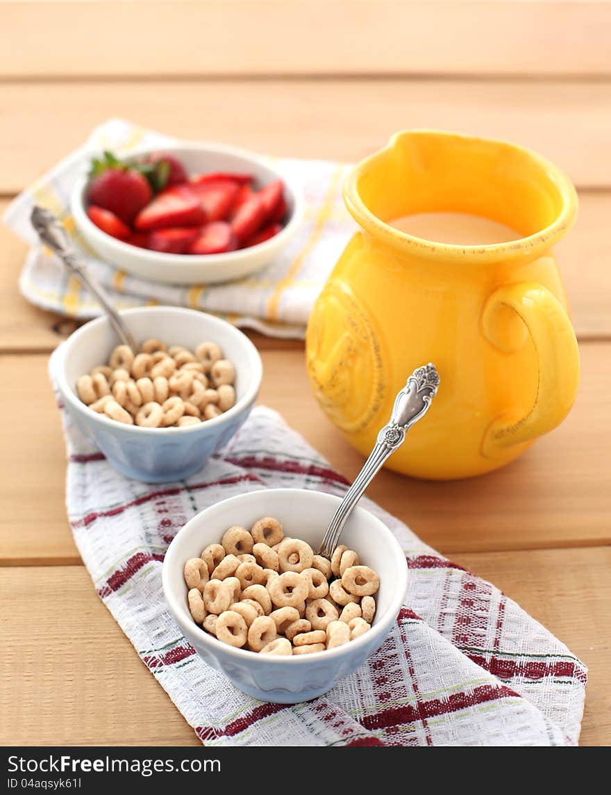 Close-up of a two bowls of cereal with a pitcher of fresh milk and strawberries.  Served on a wooden table, with a metal spoon on a napkin. Close-up of a two bowls of cereal with a pitcher of fresh milk and strawberries.  Served on a wooden table, with a metal spoon on a napkin.