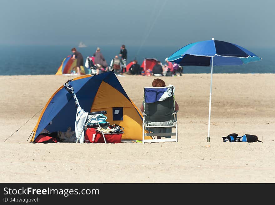 Scene at the beach with a unrecognizable person sitting on a deckchair, with a tent and a sunshade and other people in the farer background