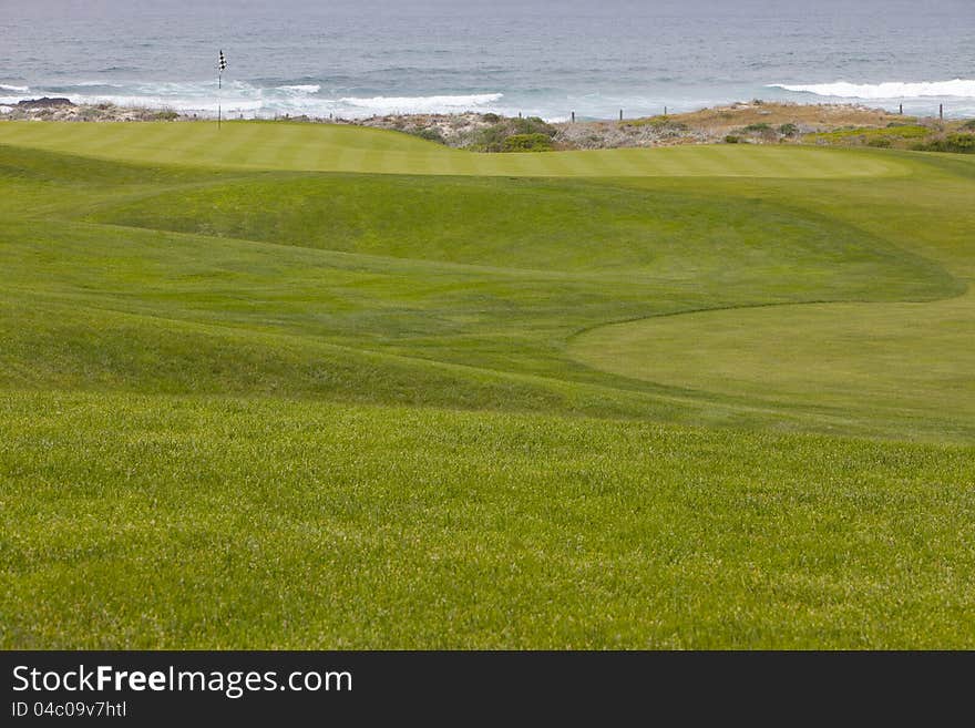 Beautiful, manicured golf course greens on a softly rolling hilly topography leading to the hole. The putting surface is located right by the ocean, with small waves washing into the shore. Beautiful, manicured golf course greens on a softly rolling hilly topography leading to the hole. The putting surface is located right by the ocean, with small waves washing into the shore.