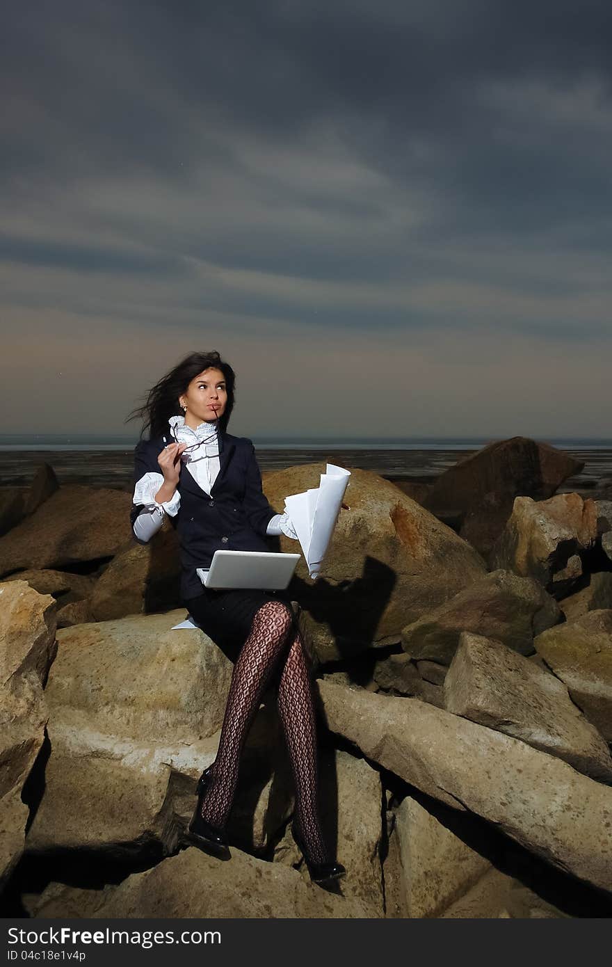 Business lady sitting on the rocks by the sea, against the backdrop of a cloudy sky. In the hands of her notebook and paper. Wind waving her hair.