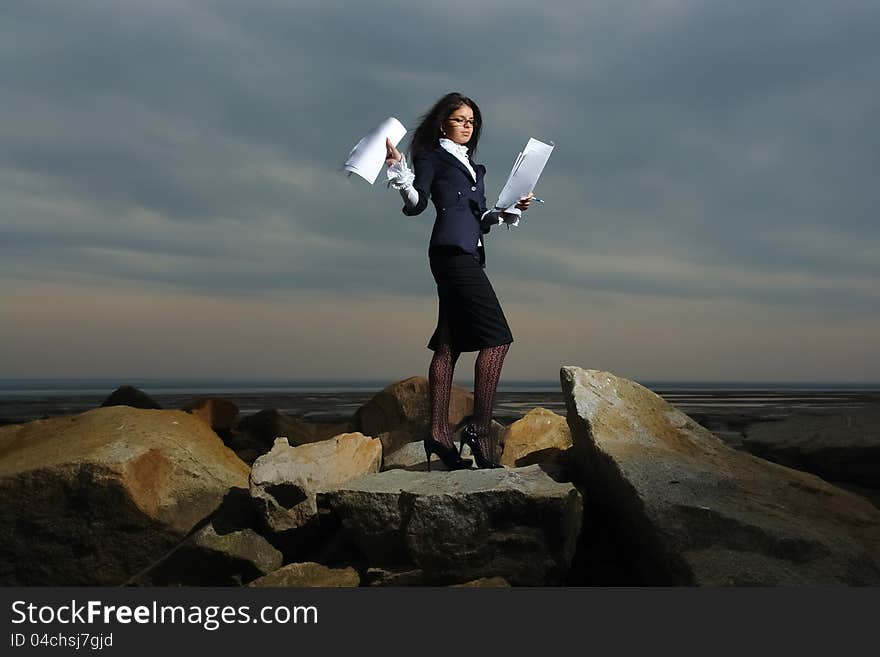 Business ladies standing on rocks by the sea, against the backdrop of a cloudy sky. In the hands of her paper. Wind waving her hair.