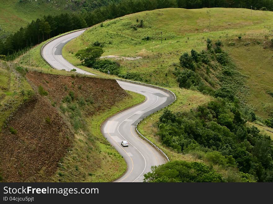 A car drives by a curvy road along the mountain scenery, surrounded by green vegetation. A car drives by a curvy road along the mountain scenery, surrounded by green vegetation.